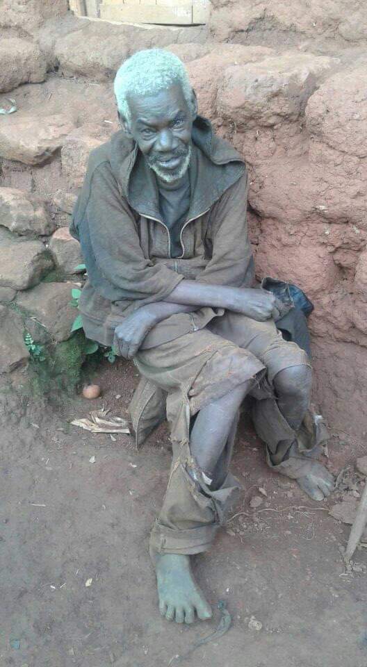 old barefoot african man sitting on a bench in front of a stone building