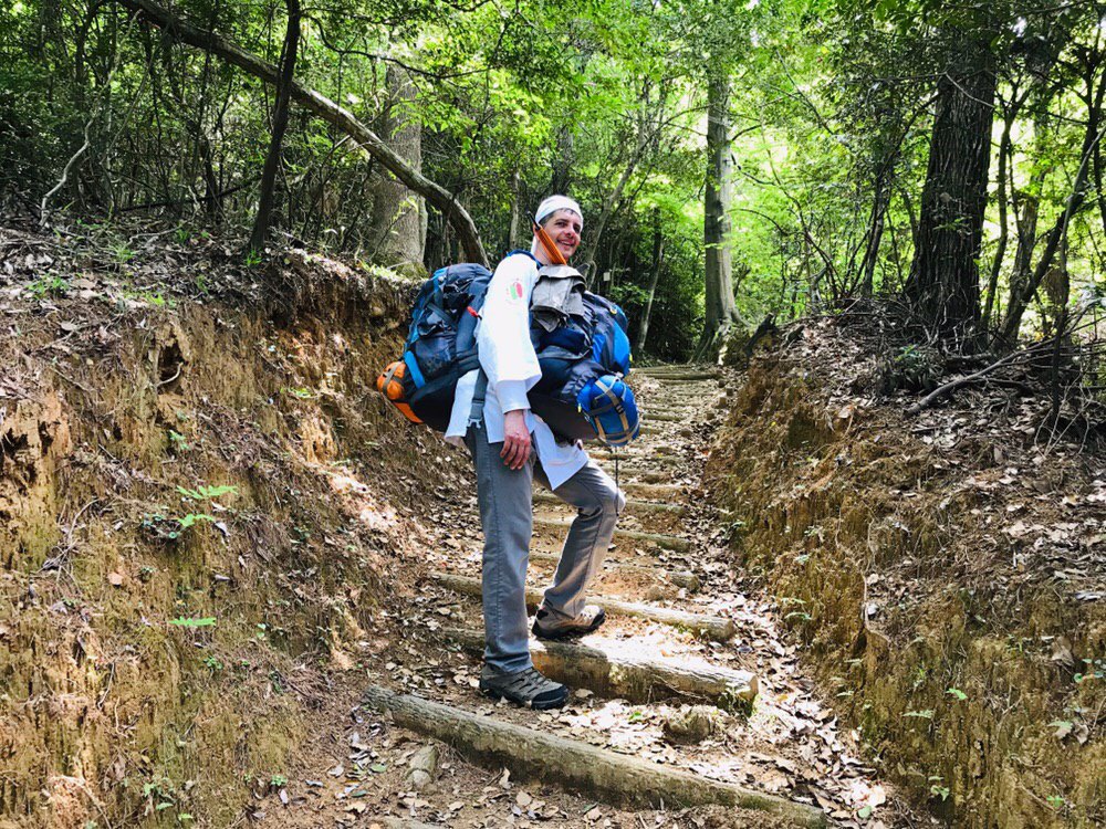 man carrying two backpacks one on his back the other on his stomach standing on steps in a forest